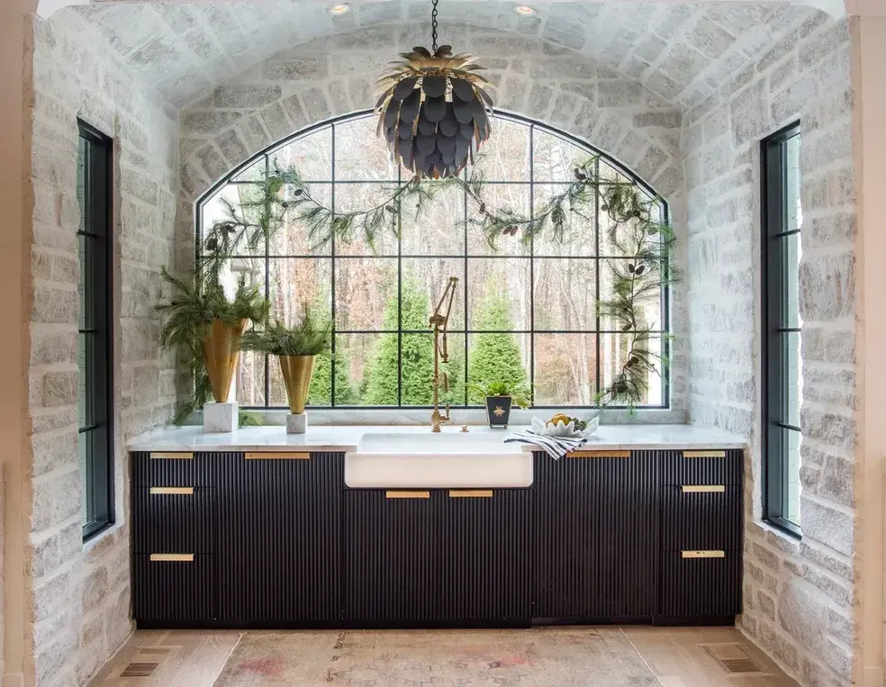 Black fluted vanity with brass hardware and an apron front sink below an arched window in a stone walled bathroom