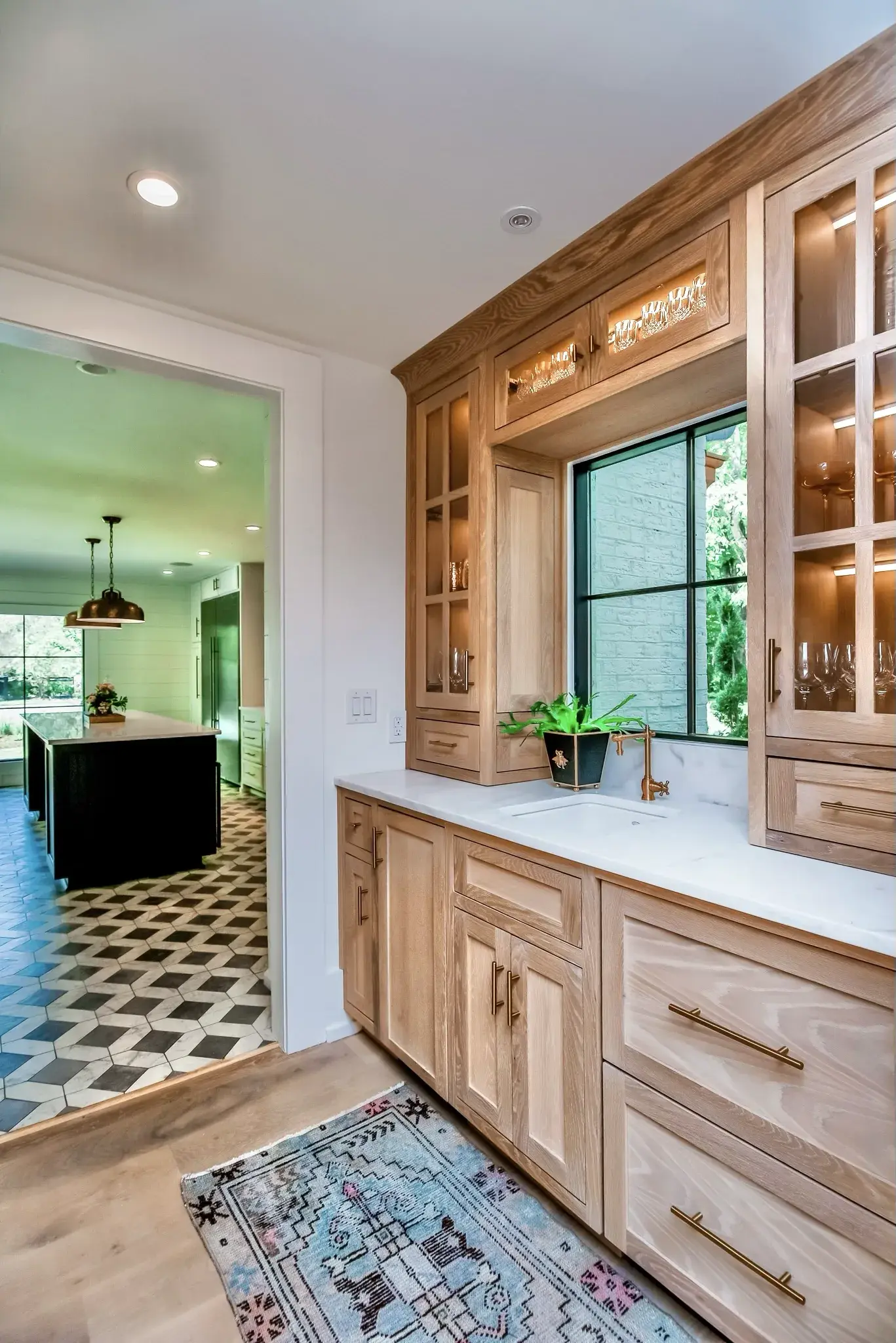 Butler pantry with white oak cabinets, lit glass fronts, a brass faucet and a view through to the kitchen