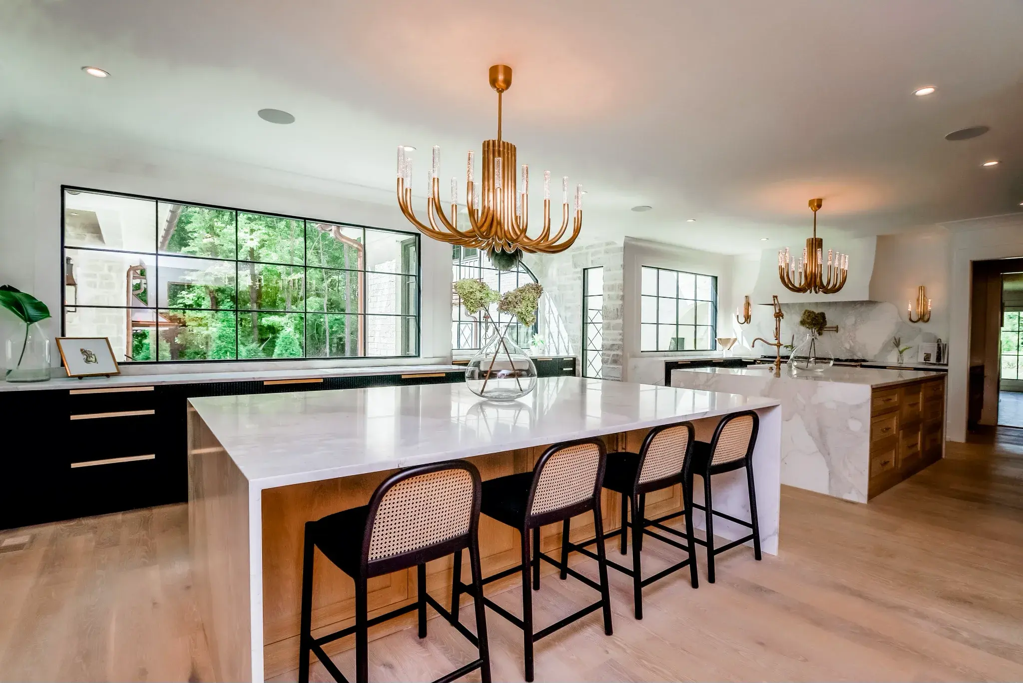 Kitchen with a marble waterfall island, cane bar stools, brass chandeliers and black steel windows