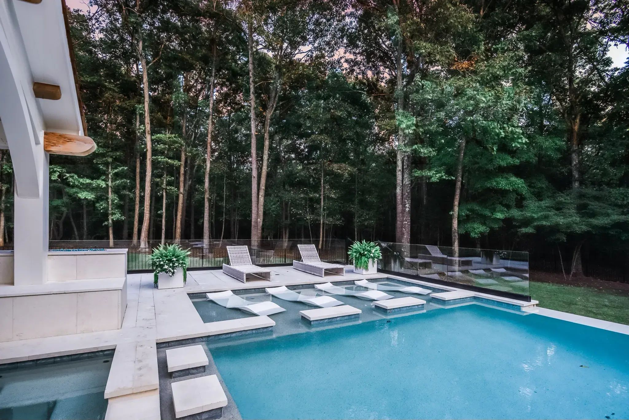 Pool with a tanning ledge, in-water lounge chairs and glass fencing backing up to woods at dusk