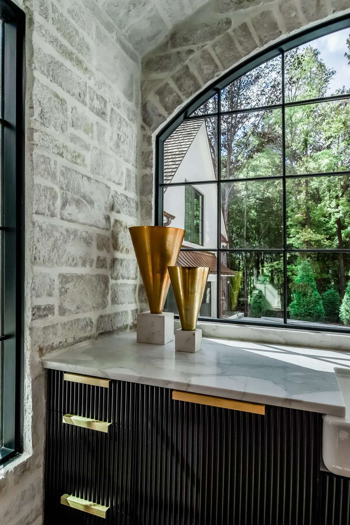 Stone walled bar with an arched steel window, black fluted cabinets with brass pulls and a marble top
